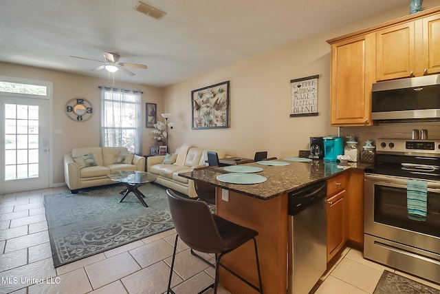 kitchen with kitchen peninsula, backsplash, ceiling fan, dark stone counters, and stainless steel appliances