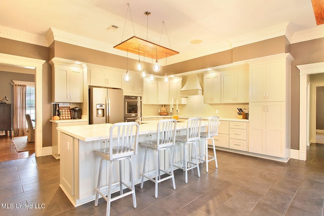 kitchen with white cabinetry, white refrigerator with ice dispenser, premium range hood, decorative light fixtures, and a center island with sink