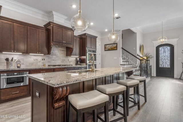 5705 Via Toscana, Ocean Springs, MS 39564 - kitchen featuring appliances with stainless steel finishes, decorative backsplash, light stone counters, a center island with sink, and light wood finished floors