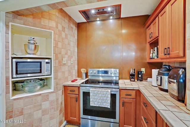 kitchen with appliances with stainless steel finishes, tile walls, tile counters, and brown cabinets