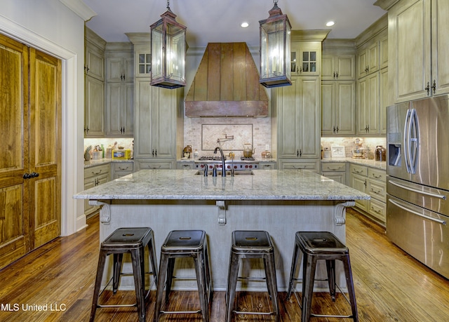 kitchen featuring stainless steel fridge, premium range hood, light stone countertops, an island with sink, and decorative backsplash