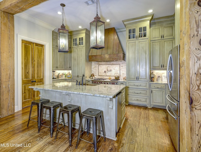 kitchen with stainless steel refrigerator, light stone counters, custom range hood, an island with sink, and decorative light fixtures