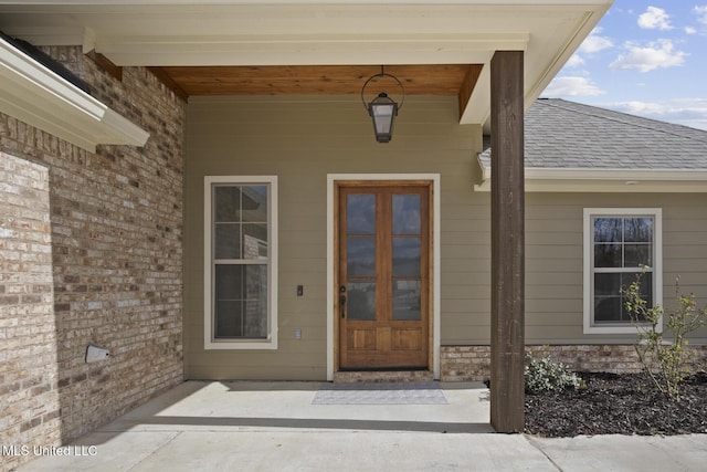 view of exterior entry with covered porch, a shingled roof, and brick siding