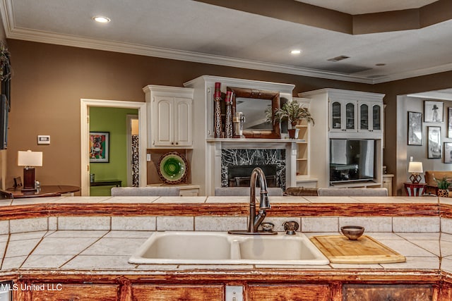 kitchen with a premium fireplace, white cabinetry, ornamental molding, tile counters, and sink