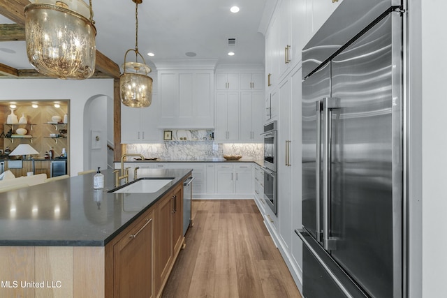 kitchen featuring arched walkways, decorative light fixtures, white cabinets, an island with sink, and stainless steel built in refrigerator