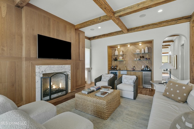 living room with coffered ceiling, light wood-style flooring, a premium fireplace, indoor wet bar, and beam ceiling