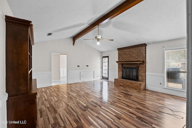 unfurnished living room with lofted ceiling with beams, a brick fireplace, a textured ceiling, ceiling fan, and light hardwood / wood-style floors