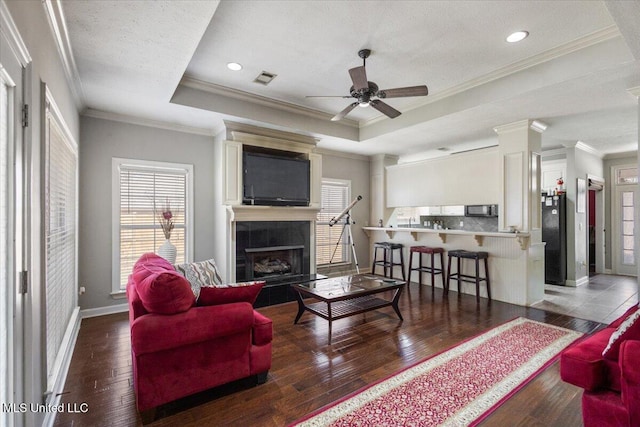 living room with dark wood-type flooring, ornamental molding, a tray ceiling, ceiling fan, and a fireplace