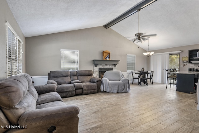 living area with lofted ceiling with beams, light wood finished floors, a textured ceiling, a stone fireplace, and ceiling fan with notable chandelier
