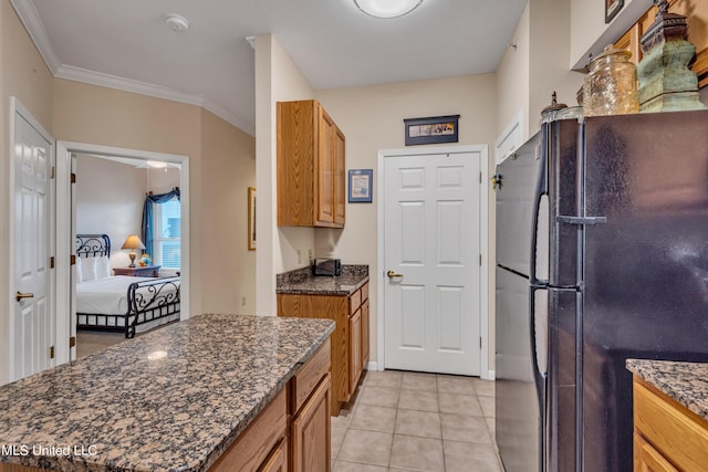 kitchen featuring freestanding refrigerator, ornamental molding, brown cabinets, dark stone countertops, and light tile patterned floors