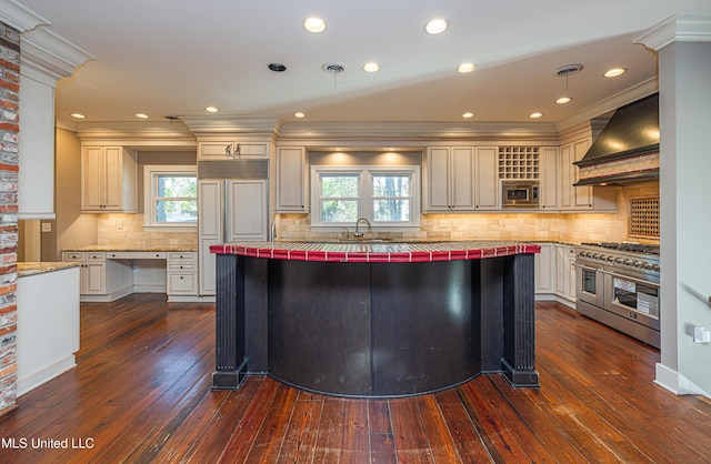 kitchen with built in appliances, tile countertops, a sink, wall chimney range hood, and dark wood finished floors