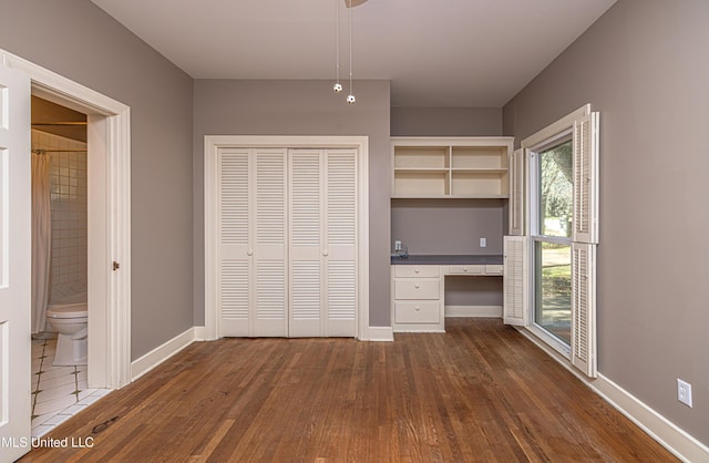 unfurnished bedroom with dark wood finished floors, baseboards, a closet, built in desk, and ensuite bath