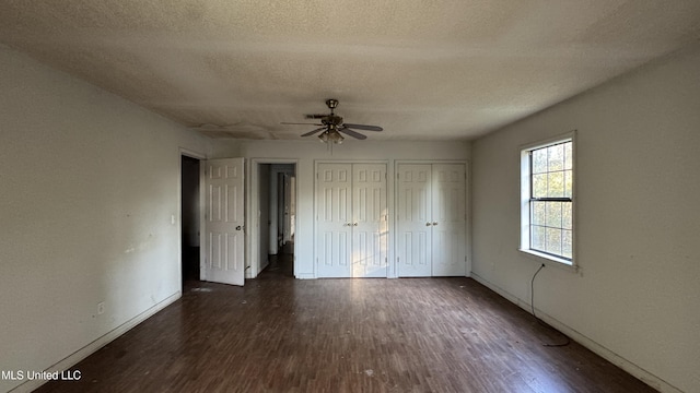 unfurnished bedroom with a textured ceiling, ceiling fan, dark wood-type flooring, and two closets