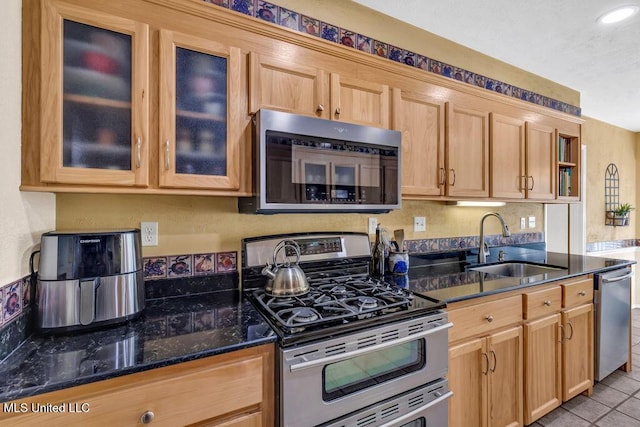 kitchen featuring light tile patterned floors, dark stone counters, glass insert cabinets, appliances with stainless steel finishes, and a sink