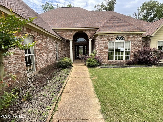doorway to property featuring brick siding, roof with shingles, and a lawn