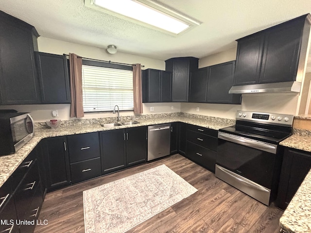 kitchen with dark wood-style floors, a sink, dark cabinetry, under cabinet range hood, and appliances with stainless steel finishes