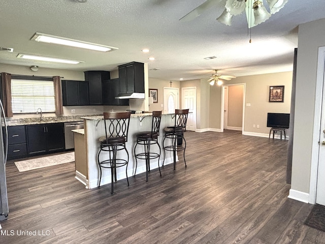 kitchen featuring stainless steel dishwasher, dark cabinetry, dark wood-type flooring, a breakfast bar area, and ceiling fan