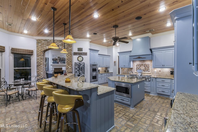 kitchen featuring premium range hood, a kitchen island with sink, hanging light fixtures, a fireplace, and stainless steel appliances