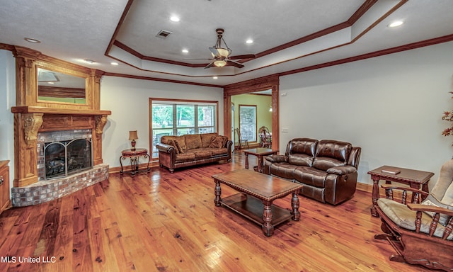 living room with crown molding, a tray ceiling, a brick fireplace, and light wood-type flooring