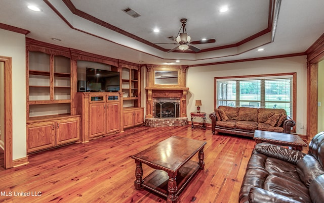 living room featuring ceiling fan, a tray ceiling, a brick fireplace, light hardwood / wood-style flooring, and ornamental molding