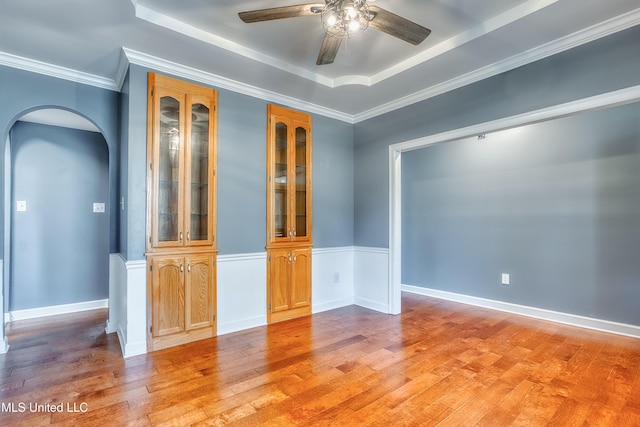 empty room with a raised ceiling, arched walkways, ceiling fan, light wood-style floors, and crown molding