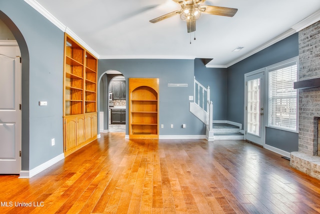unfurnished living room with arched walkways, stairway, light wood-style flooring, a ceiling fan, and ornamental molding