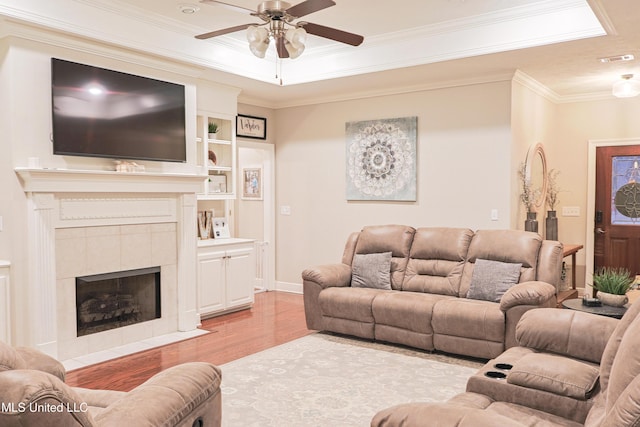 living room with crown molding, light hardwood / wood-style flooring, a raised ceiling, a tile fireplace, and ceiling fan