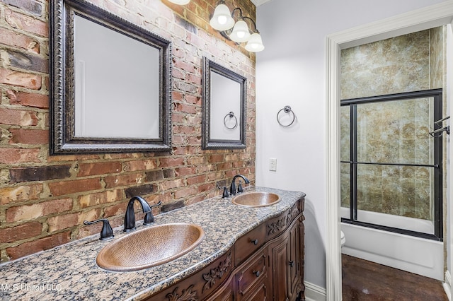 full bath featuring shower / bath combination with glass door, double vanity, brick wall, and a sink