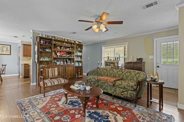 living room featuring a textured ceiling, ornamental molding, a ceiling fan, visible vents, and wood-type flooring