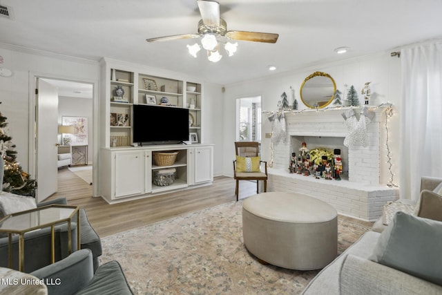 living room with ceiling fan, a brick fireplace, built in features, light hardwood / wood-style floors, and ornamental molding