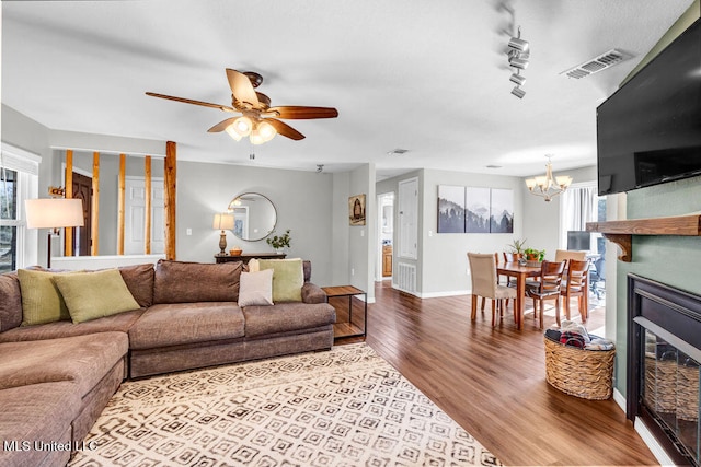 living area with visible vents, a glass covered fireplace, wood finished floors, baseboards, and ceiling fan with notable chandelier