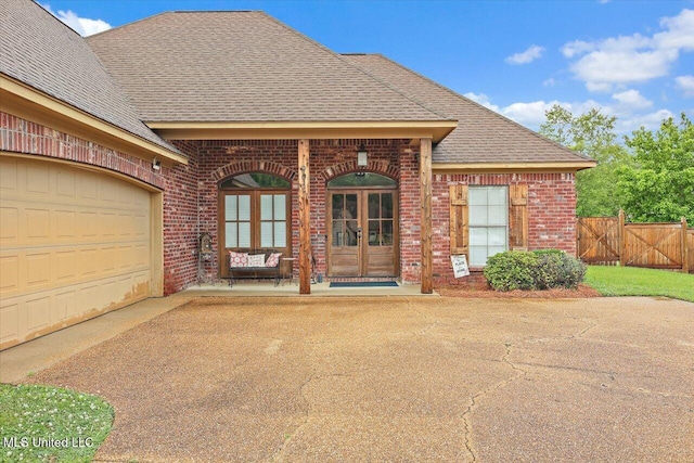 view of exterior entry with a garage, a shingled roof, driveway, brick siding, and french doors