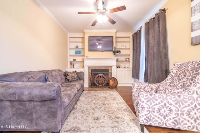 living room featuring a tiled fireplace, wood-type flooring, crown molding, built in shelves, and ceiling fan