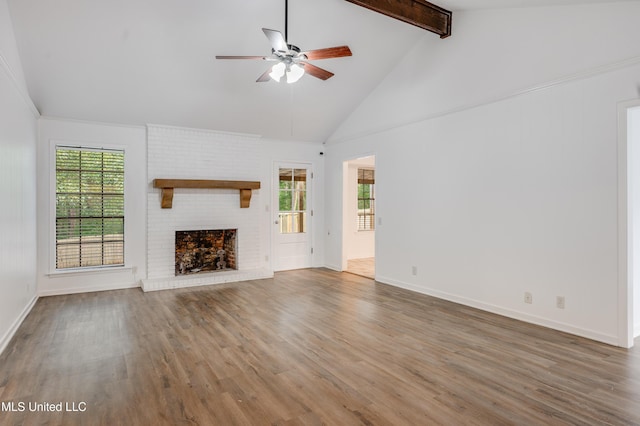 unfurnished living room with a ceiling fan, healthy amount of natural light, beamed ceiling, wood finished floors, and a brick fireplace