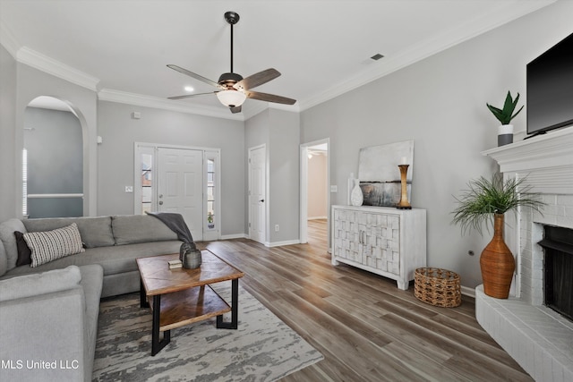 living area with a brick fireplace, a ceiling fan, wood finished floors, visible vents, and ornamental molding