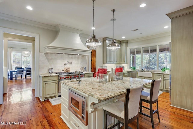 kitchen with light stone countertops, light wood-type flooring, custom exhaust hood, stainless steel appliances, and a kitchen island with sink