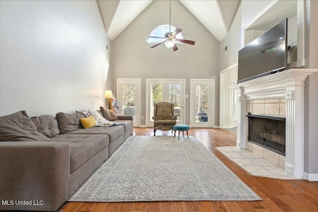 living room with ceiling fan, a tiled fireplace, high vaulted ceiling, and light hardwood / wood-style flooring