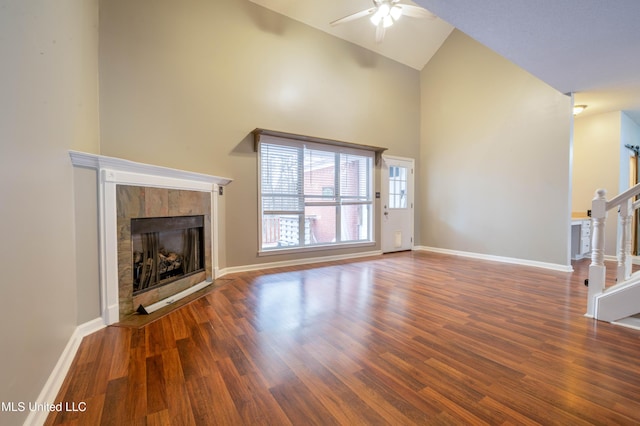 unfurnished living room featuring a tiled fireplace, wood finished floors, high vaulted ceiling, baseboards, and stairs