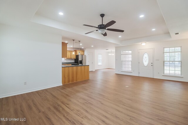 unfurnished living room with light wood finished floors, visible vents, a tray ceiling, a healthy amount of sunlight, and ceiling fan with notable chandelier