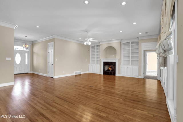 unfurnished living room with ceiling fan with notable chandelier, built in shelves, hardwood / wood-style flooring, crown molding, and a tiled fireplace