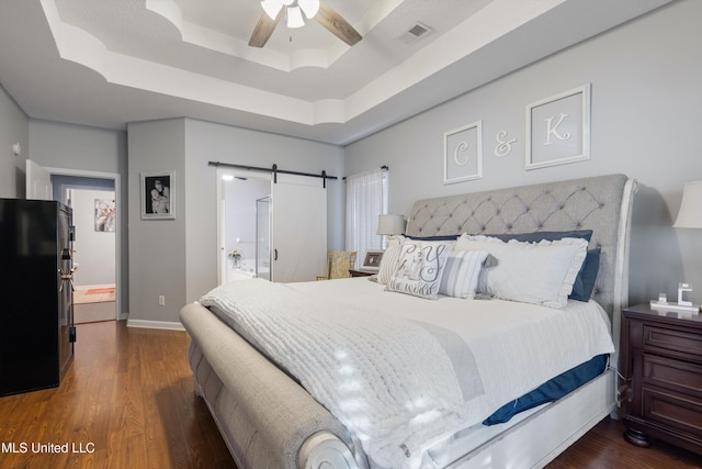 bedroom featuring a barn door, dark hardwood / wood-style floors, ceiling fan, and a tray ceiling