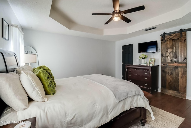 bedroom with dark wood-style flooring, a raised ceiling, visible vents, a barn door, and ceiling fan