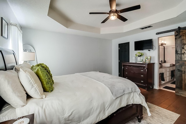 bedroom with a barn door, visible vents, ceiling fan, dark wood-style flooring, and a tray ceiling