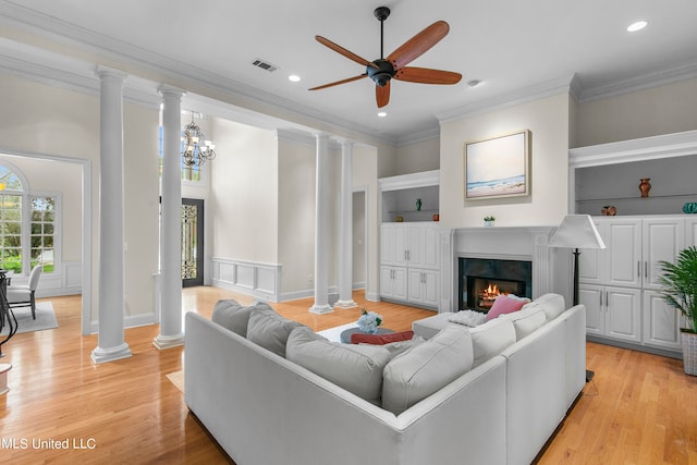 living room with crown molding, decorative columns, light wood-type flooring, built in shelves, and ceiling fan with notable chandelier