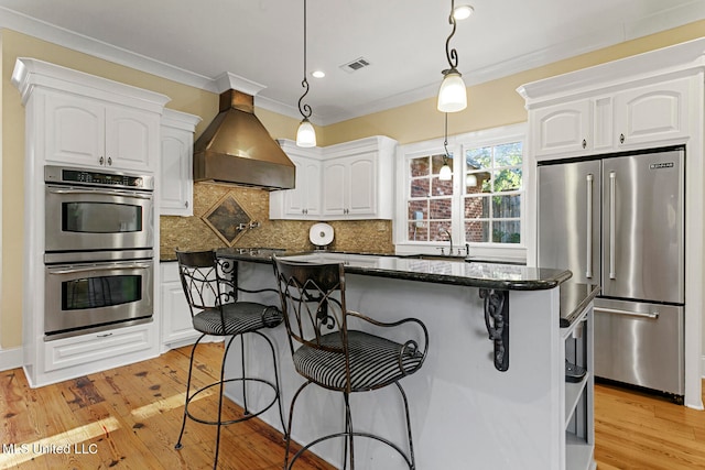 kitchen featuring wall chimney range hood, light hardwood / wood-style flooring, white cabinets, decorative light fixtures, and appliances with stainless steel finishes