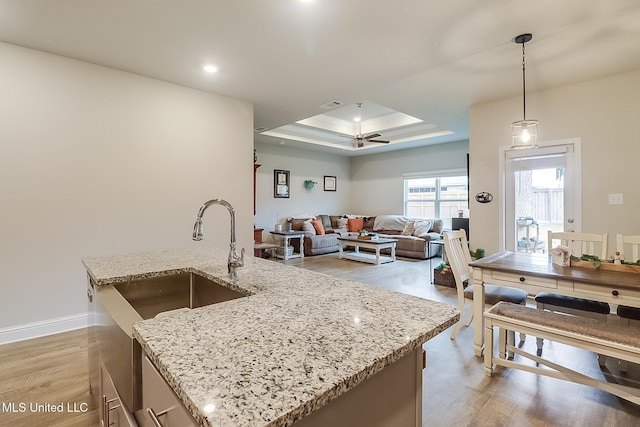 kitchen with hanging light fixtures, an island with sink, a raised ceiling, and light stone counters