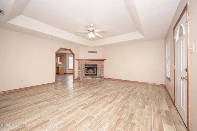 unfurnished living room with ceiling fan, a tray ceiling, a brick fireplace, and light wood-type flooring