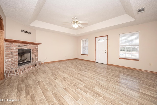 unfurnished living room featuring a brick fireplace, a tray ceiling, a textured ceiling, and ceiling fan