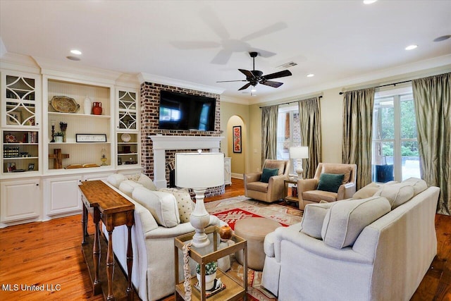 living room with ornamental molding, light wood-style flooring, a ceiling fan, a brick fireplace, and visible vents