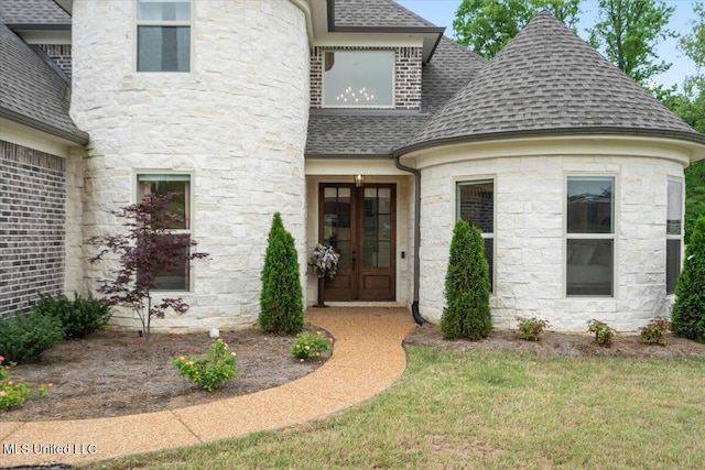 entrance to property featuring a shingled roof, french doors, stone siding, and a lawn